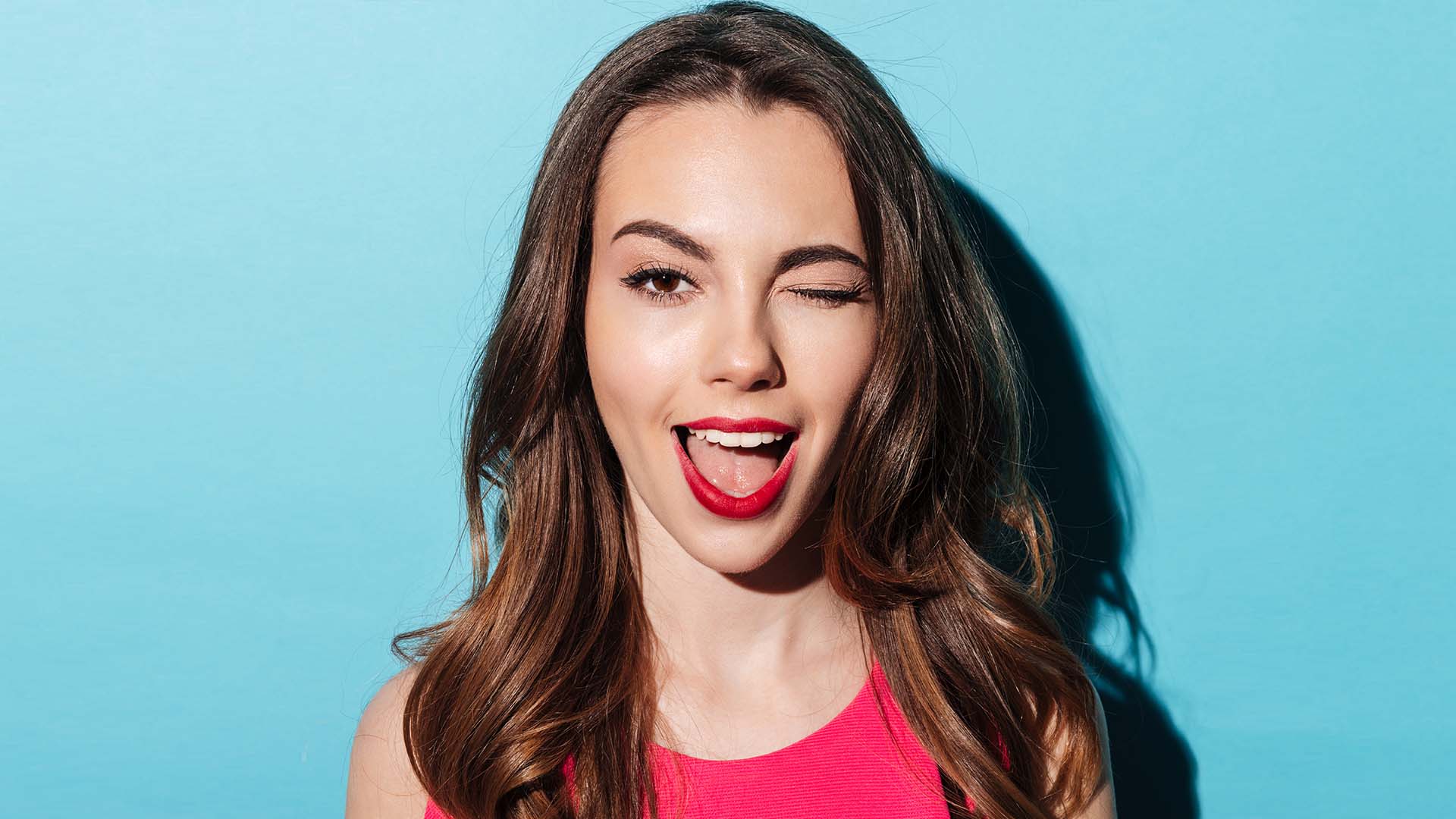 Brunette woman in hot pink shirt and lipstick, winking and smiling. Light blue background.